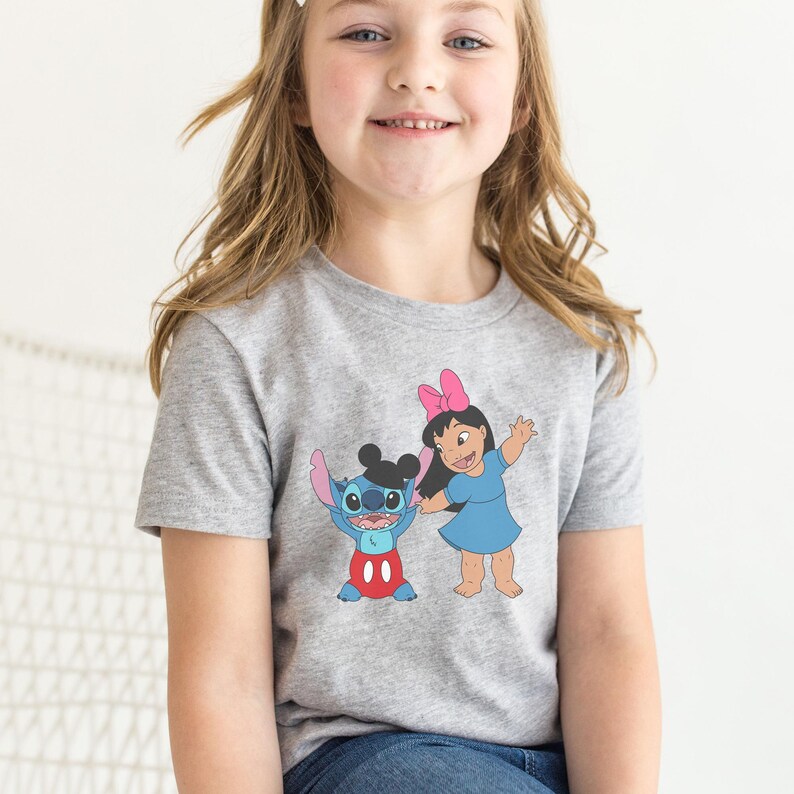 A young girl wearing a gray t-shirt with a graphic of two cartoon characters, Stitch and Lilo, smiling and waving at the camera.