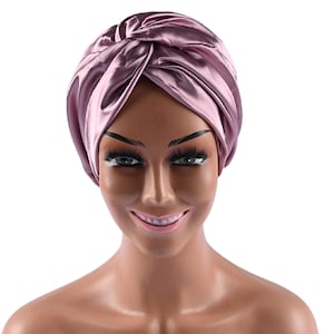 May include: A mauve satin turban is displayed on a mannequin. The turban is knotted at the top and has a shiny, smooth texture. The mannequin has dark eyebrows, long eyelashes, and a slight smile. The background is white.