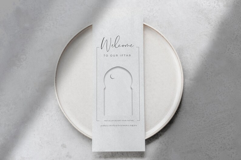 May include: A white card with the text "Welcome to our Iftar" and a decorative archway with a crescent moon. The card is on a white plate.