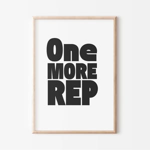 May include: A framed print with the words "One MORE REP" in bold, black letters. The text is centered on a white background, and the frame is a light wood color. The print is a motivational piece.