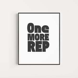 May include: A framed print with the words "One MORE REP" in bold, black letters on a white background. The print is in a black frame and is displayed against a white wall.