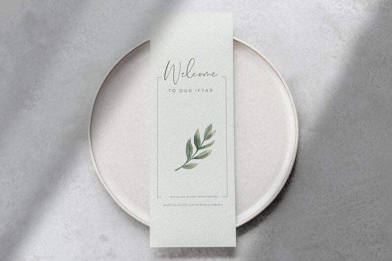 K&ouml;nnte beinhalten: Eine wei&szlig;e Karte mit dem Text "Willkommen zu unserem Iftar" und einer gr&uuml;nen Blatt-Illustration. Die Karte liegt auf einem wei&szlig;en Teller.