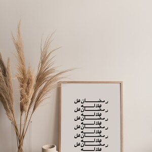 May include: A framed print with the Arabic text "Ramadan هل هلاله" repeated multiple times. The print is in a light wood frame and is hanging on a white wall. There are dried pampas grasses in a clear vase to the left of the print.