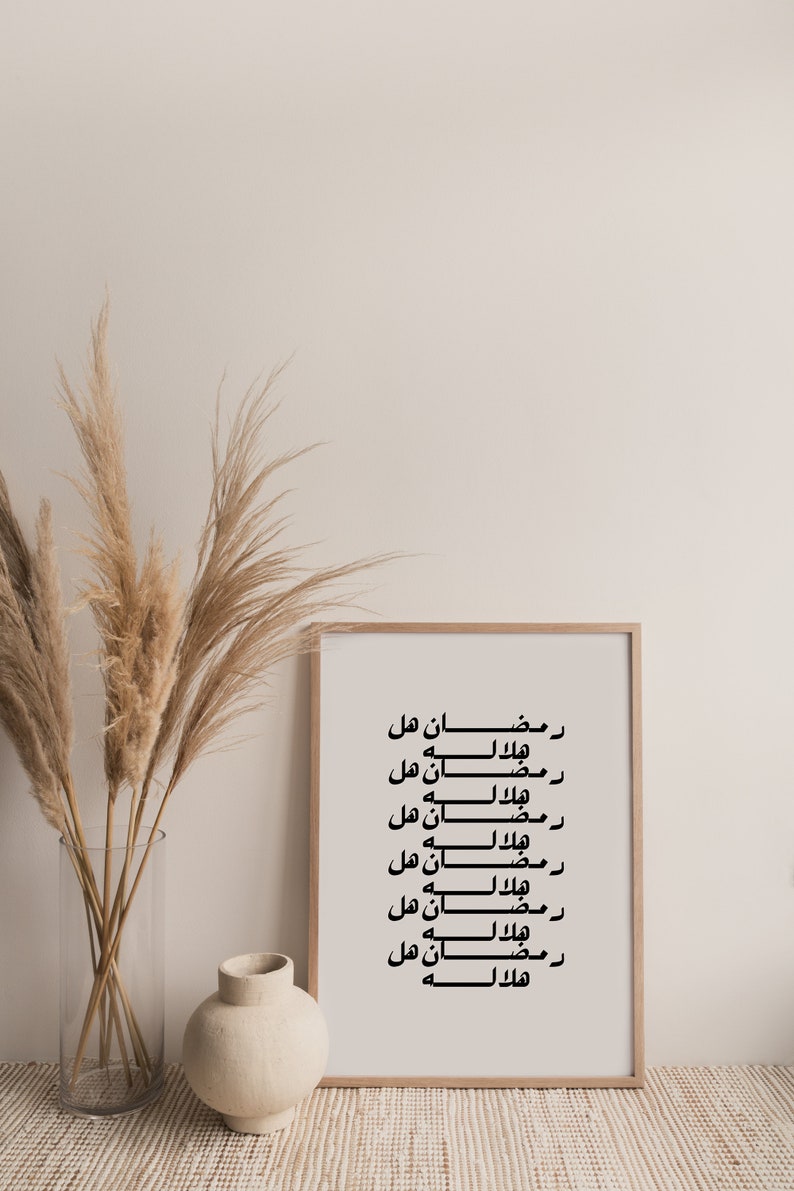 May include: A framed print with the Arabic text "Ramadan هل هلاله" repeated multiple times. The print is in a light wood frame and is hanging on a white wall. There are dried pampas grasses in a clear vase to the left of the print.