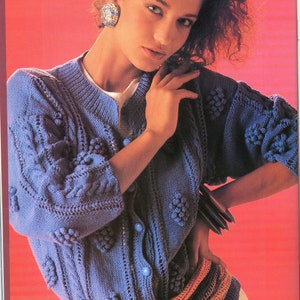 May include: A blue knitted cardigan with a cable knit pattern and decorative bobbles. The cardigan has a button closure and is worn with a brown and orange belt.