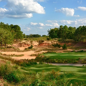 Może przedstawiać: Pole golfowe z zielonym fairwayem i bunkrami piaskowymi. Pole jest otoczone drzewami i błękitnym niebem z białymi chmurami.