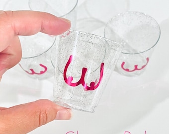 Könnte beinhalten: Ein Set aus durchsichtigen Kunststoff-Schnapsgläsern mit silbernem Glitzer-Finish und einer leuchtend pinken Chrom-Grafik, die eine stilisierte Brustkontur zeigt. Der Text Chrome Pink ist unten in Pink gedruckt und benennt die Farbe.