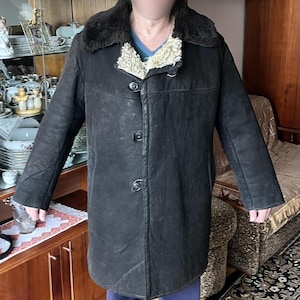 Pode incluir: Um casaco de couro marrom com gola de pele. O casaco tem fecho de botões e é forrado com tecido marrom. A pessoa que veste o casaco está em pé em uma sala com armários de madeira e um tapete.