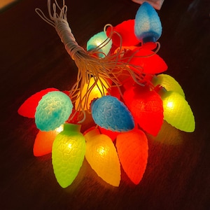 May include: A string of Christmas lights with red, blue, green and yellow bulbs. The lights are in the shape of a pine cone and are strung together with white wire.