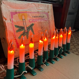 Könnte beinhalten: Eine Lichterkette mit vintage Weihnachtslichtern mit warmem orangefarbenem Schein. Die Lichter sind kerzenförmig und haben grüne Klammern. Die Schachtel für die Lichter ist im Hintergrund sichtbar und trägt die Aufschrift "Гирлянда "Сюрприз" (Girlyanda "Surprise") in russischer Sprache.