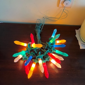 Può includere: Una ghirlanda di luci natalizie colorate a forma di candela, con un filo verde. Le luci sono collegate a una presa elettrica e sono accese.