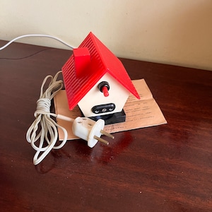 Puede incluir: Una toma de corriente de forma de casa roja y blanca con un cable y un enchufe blancos. La toma tiene un techo rojo y un interruptor negro. El texto en el papel junto a la toma dice "ВНИМАНИЕ!"