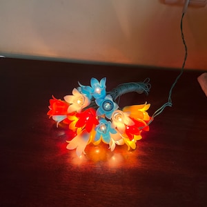 May include: A string of fairy lights shaped like flowers in blue, white, orange, and red. The lights are plugged into an outlet and are lit.