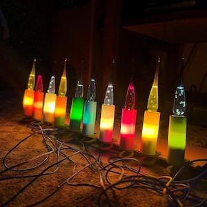 Könnte beinhalten: Eine Lichterkette aus zehn elektrischen Kerzen mit farbigen Sockeln und klaren, spitz zulaufenden Glühbirnen. Jede Kerze strahlt ein warmes Licht in verschiedenen Farben wie Rot, Gelb, Grün, Blau und Weiß aus und ist mit einem dünnen schwarzen Kabel verbunden.