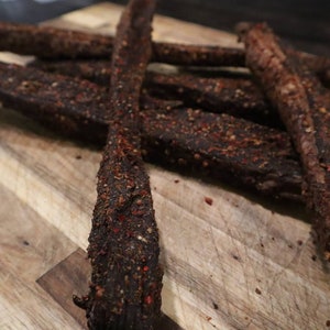 May include: Close-up of several pieces of dried meat, likely beef jerky, arranged on a wooden cutting board. The meat is dark brown with a reddish spice coating, and the board has a light brown, striped pattern.