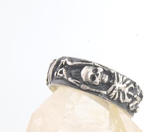 May include: A silver-toned ring featuring a detailed skull and skeleton design. The ring is set against a white background, showcasing the intricate carvings and gothic aesthetic. The ring's design includes a skull and crossbones.