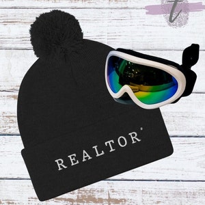 May include: Black beanie with a white pom pom and the word "REALTOR" embroidered on the front. The beanie is paired with white ski goggles with a rainbow iridescent lens.