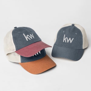 May include: Two trucker hats with a white mesh back and a blue denim front. The front of the hats have the letters "KW" embroidered in white. One hat has a burgundy brim and the other has a brown brim.