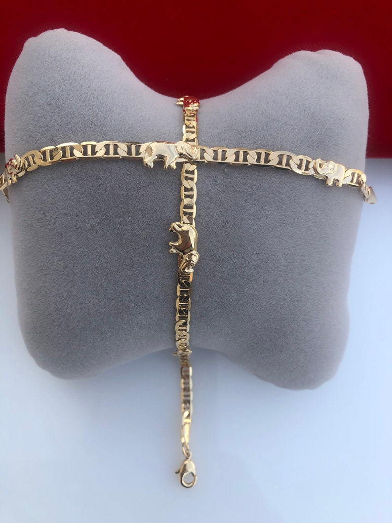 Peut inclure: Un bracelet dor&eacute; avec un motif de breloques &eacute;l&eacute;phants. Le bracelet est compos&eacute; de petits maillons interconnect&eacute;s et est pr&eacute;sent&eacute; sur un coussin gris. Les breloques &eacute;l&eacute;phants sont espac&eacute;es uniform&eacute;ment.