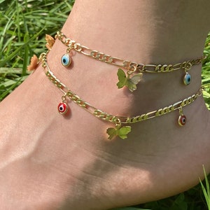 May include: Gold-colored anklets with butterfly and evil eye charms. The anklets are made of a chain link design. One anklet has a red evil eye charm, and the other has a blue evil eye charm. The background is green grass.