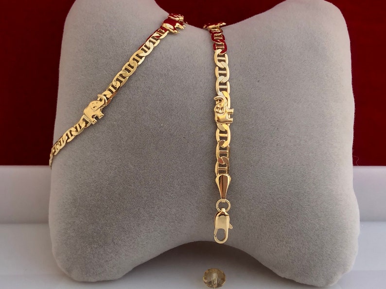 Peut inclure: Bracelet en cha&icirc;ne de couleur or avec un charm en forme d'&eacute;l&eacute;phant. Le bracelet pr&eacute;sente un design de cha&icirc;ne nautique et une fermeture &agrave; fermoir homard. Le charm en forme d'&eacute;l&eacute;phant est visible sur le bracelet. Fond gris et rouge.