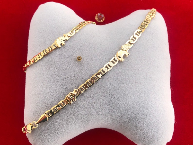 Peut inclure: Un bracelet dor&eacute; avec un charm en forme d'&eacute;l&eacute;phant et un motif de cha&icirc;ne nautique. Le bracelet est pr&eacute;sent&eacute; sur un coussin blanc sur fond rouge. Le bracelet est dot&eacute; d'une fermeture &agrave; fermoir homard.