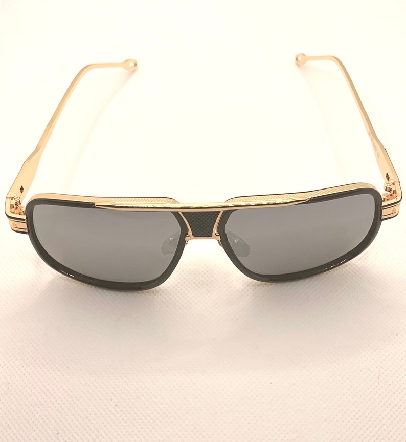 May include: Gold-framed sunglasses with gray lenses. The sunglasses have a black bridge and black accents on the frame. The temples are thin and gold, with a small diamond-shaped detail near the hinges. The lenses are a dark gray color.