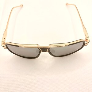 May include: Gold-framed sunglasses with dark gray lenses. The sunglasses have a black bridge and temples, and a textured design on the frame. The arms are thin and gold-colored.