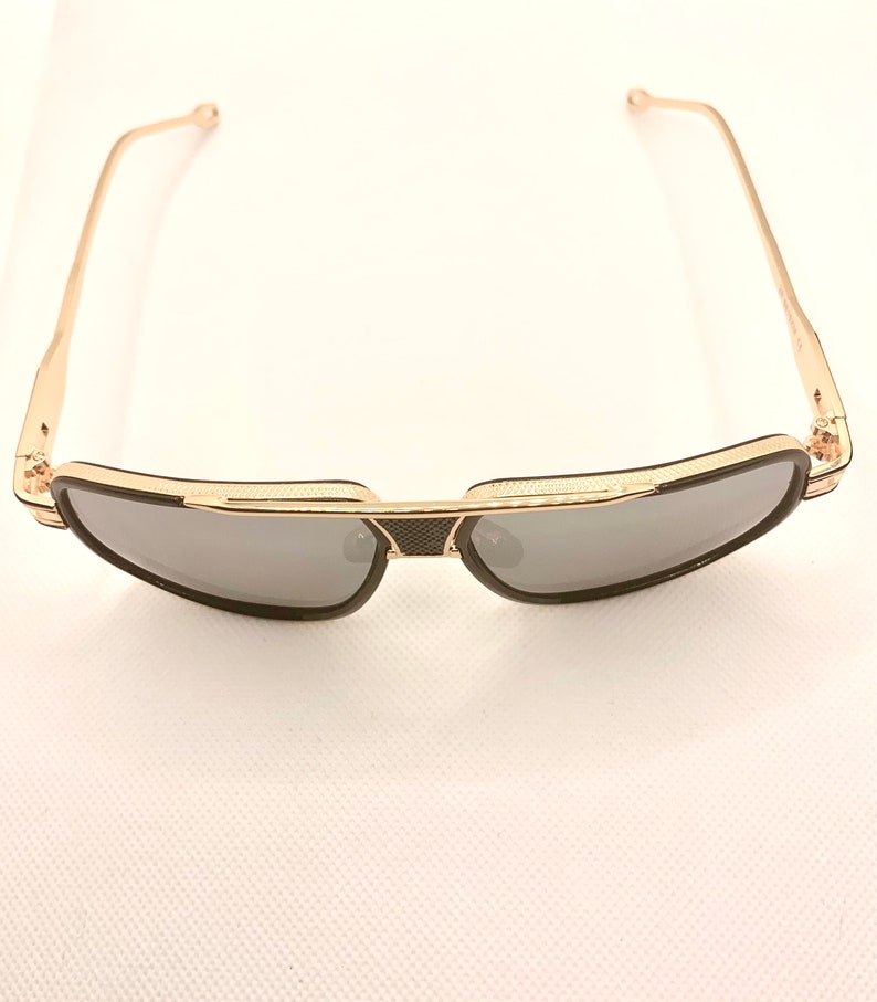May include: Gold-framed sunglasses with dark gray lenses. The sunglasses have a black bridge and temples, and a textured design on the frame. The arms are thin and gold-colored.
