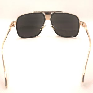 May include: Gold-framed sunglasses with dark lenses. The sunglasses have a square shape with a gold bridge and temples. The lenses are a dark gray color, and the frames have a black border.