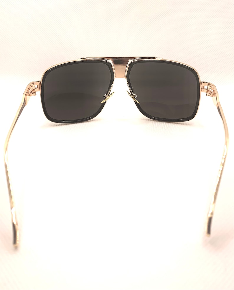 May include: Gold-framed sunglasses with dark lenses. The sunglasses have a square shape with a gold bridge and temples. The lenses are a dark gray color, and the frames have a black border.