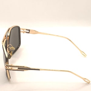 May include: Gold-framed sunglasses with dark lenses. The sunglasses have a rectangular shape and a double bridge. The temples are thin and curve slightly. The overall design is sleek and modern.