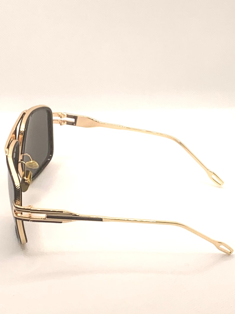 May include: Gold-framed sunglasses with dark lenses. The sunglasses have a rectangular shape and a double bridge. The temples are thin and curve slightly. The overall design is sleek and modern.