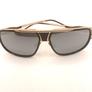 May include: Gold-framed sunglasses with dark gray lenses. The sunglasses have a double bridge design and black accents. The temples are thin and gold. The lenses are a rounded rectangular shape. The text "JR 661-" is visible on the frame.