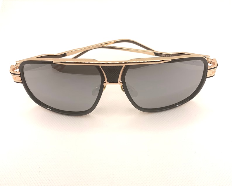 May include: Gold-framed sunglasses with dark gray lenses. The sunglasses have a double bridge design and black accents. The temples are thin and gold. The lenses are a rounded rectangular shape. The text "JR 661-" is visible on the frame.