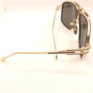 May include: Gold-framed sunglasses with dark lenses. The sunglasses have a square shape and a textured pattern on the frame. The arms are thin and gold, with a black accent near the hinges. The lenses are a dark gray color.