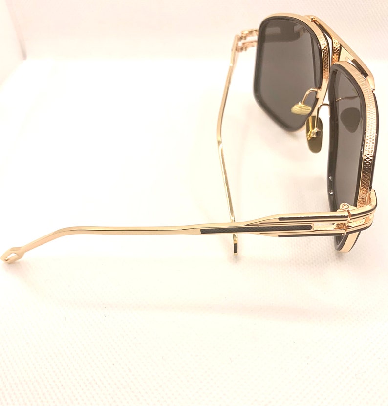 May include: Gold-framed sunglasses with dark lenses. The sunglasses have a square shape and a textured pattern on the frame. The arms are thin and gold, with a black accent near the hinges. The lenses are a dark gray color.