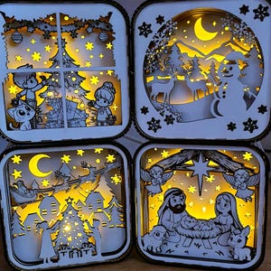 May include: Four illuminated Christmas-themed shadow boxes. Each box features a different holiday scene: children decorating a tree, a snowman with deer, Santa's sleigh, and the Nativity scene. The boxes are white with a warm yellow glow.