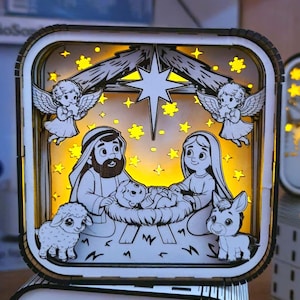 May include: A wooden, illuminated nativity scene with a square frame. The scene features a star, angels, and figures of Mary, Joseph, and baby Jesus. A lamb and donkey are also present. The background glows with yellow light.