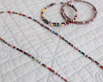 Op de afbeelding: Een kleurrijke kralenketting en twee kralenarmbanden op een witte gestikte stoffen achtergrond.