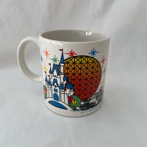 May include: White ceramic mug featuring a colorful illustration of a castle, a geometric sphere, and a boat. The design includes blue, yellow, red, and green accents, with starburst details. The mug has a curved handle.