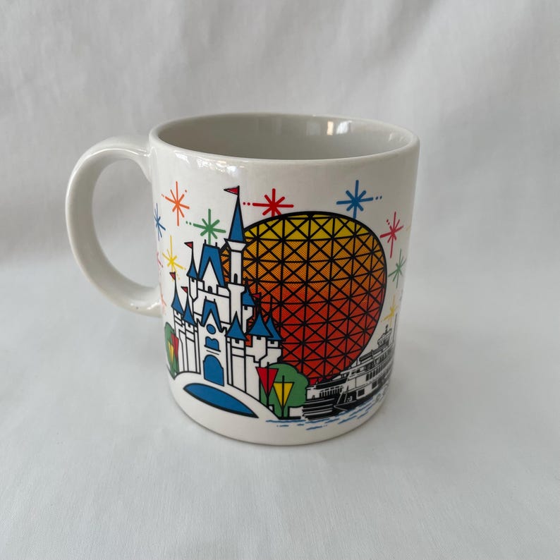 May include: White ceramic mug featuring a colorful illustration of a castle, a geometric sphere, and a boat. The design includes blue, yellow, red, and green accents, with starburst details. The mug has a curved handle.