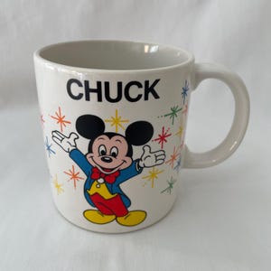 May include: White ceramic mug featuring Mickey Mouse in a blue jacket, yellow vest, and red bow tie. The mug has the name "CHUCK" in black letters and colorful starburst designs. A classic Disney collectible.