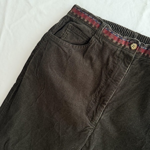 Puede incluir: Pantalones de pana marrón oscuro con diseño de cintura alta y elástica. La cintura está decorada con un ribete de patrones geométricos en rojo, azul y blanco, y cuenta con un cierre de un solo botón situado en el centro.