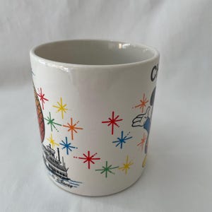 May include: White ceramic mug with colorful illustrations. Features a steamboat, a cartoon character, and starburst designs in red, yellow, blue, and green. The mug has a glossy finish and a simple, cylindrical shape.