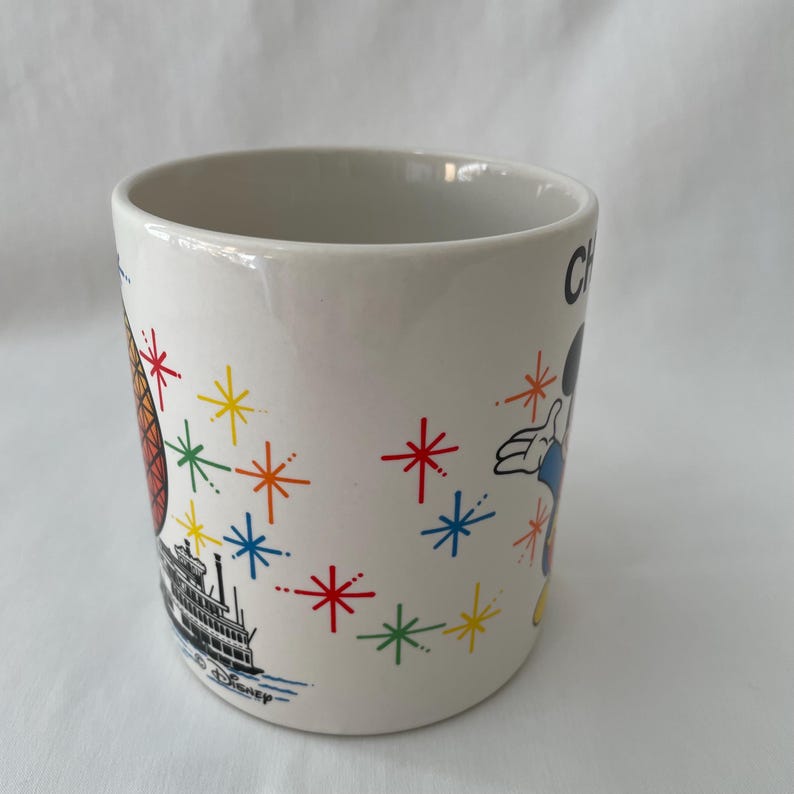 May include: White ceramic mug with colorful illustrations. Features a steamboat, a cartoon character, and starburst designs in red, yellow, blue, and green. The mug has a glossy finish and a simple, cylindrical shape.