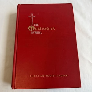 May include: A red hardcover book titled The Methodist Hymnal with a gold cross and emblem on the front cover. The text Christ Methodist Church is printed in gold near the bottom. The book has a textured finish and a vertical spine on the left side.