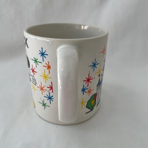 May include: White ceramic mug with a handle, featuring colorful illustrations of a cartoon character, a castle, and starbursts. The mug has a glossy finish and is suitable for hot beverages.