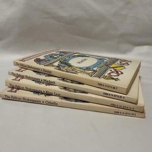 Peut inclure: Une pile de quatre livres de poche de la série The Pelican Shakespeare, comprenant Hamlet, Macbeth, King Lear et Othello. Chaque livre présente une couverture couleur crème avec des illustrations décoratives et l'ISBN imprimé sur le dos.