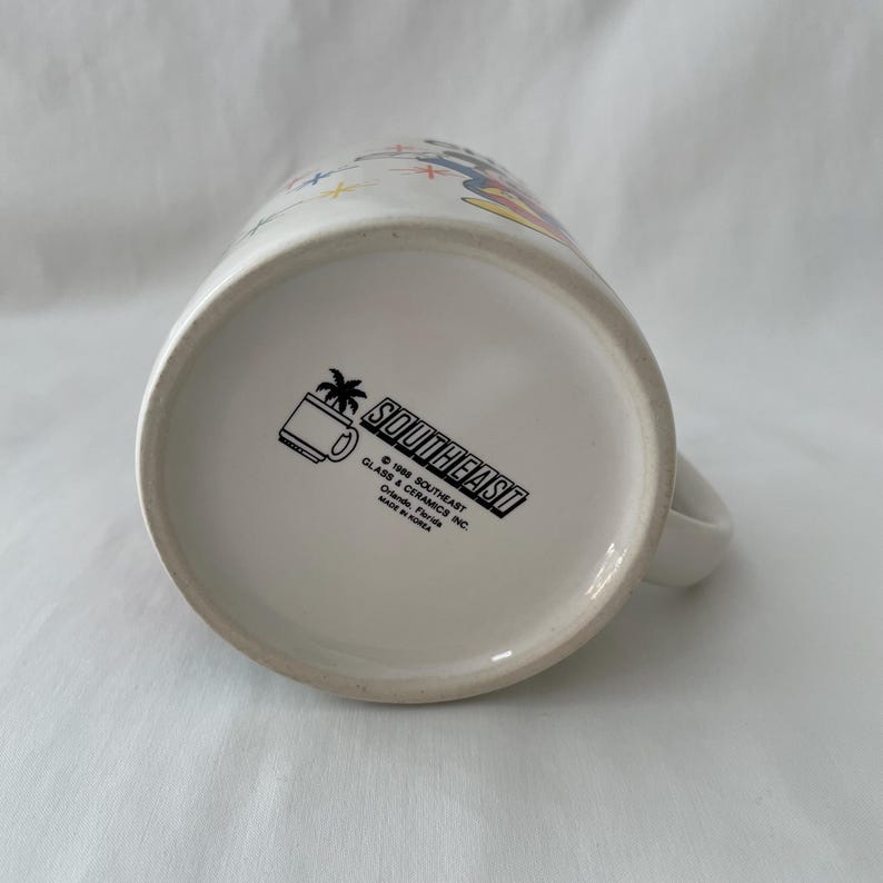 May include: White ceramic mug with a handle. The bottom of the mug features the text "SOUTHEAST" in a black rectangle, along with a palm tree and mug graphic. The mug is marked "MADE IN KOREA".